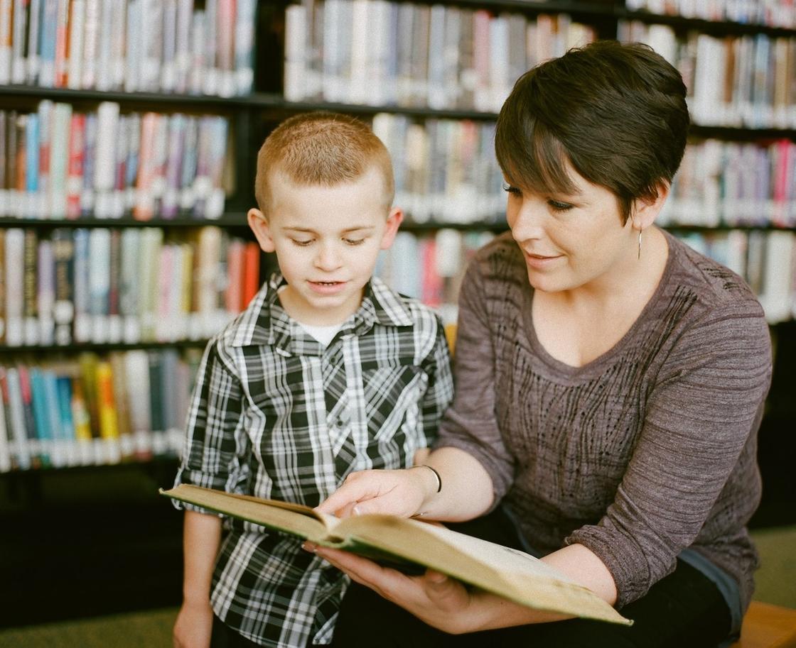 Жінка з короткою стрижкою, сидячи в бібліотеці і читає книгу хлопчика.  Хлопчик одягнений у картату сорочку