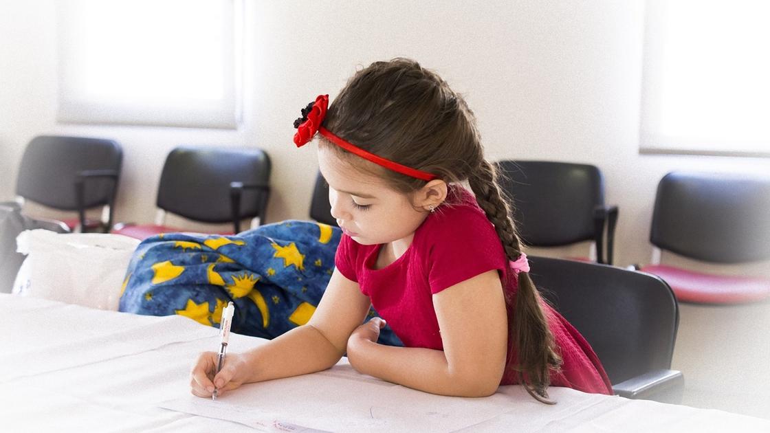 Дівчинка з довгу кіску в червоній футболці сидить за столом і пише в зошит